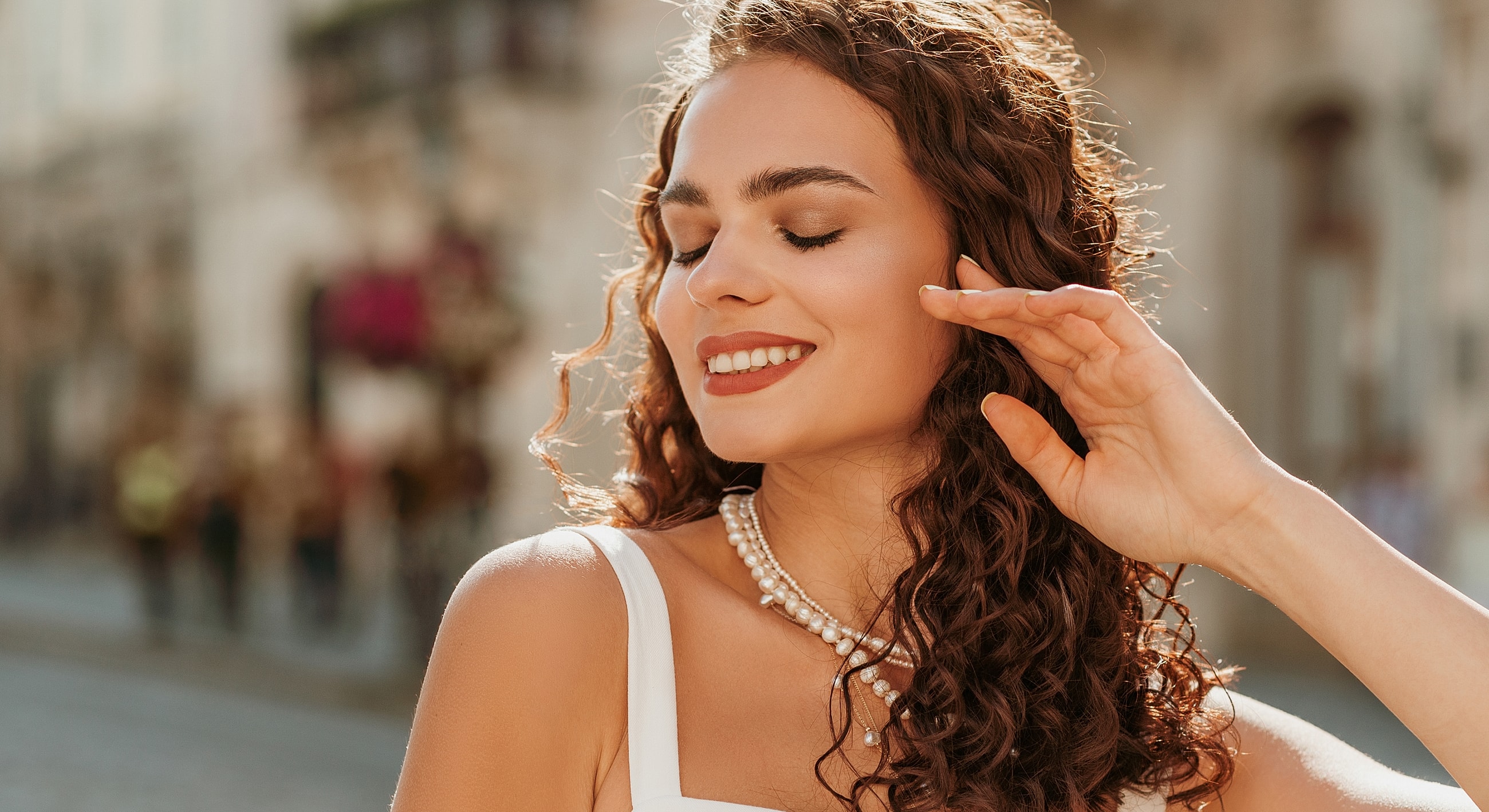 Woman smiling outdoors with curly hair and pearls.